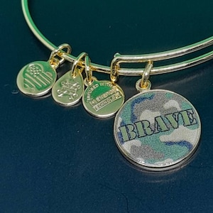 Puede incluir: Pulsera de oro con varios charms, incluyendo un charm redondo con un patrón de camuflaje y la palabra "BRAVE" en letras blancas.
