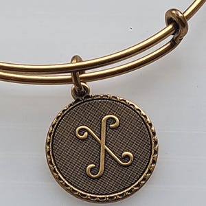 May include: Gold-toned bangle bracelet with a circular charm. The charm features a textured background and a stylized, swirling design. The bracelet has a simple, adjustable design.