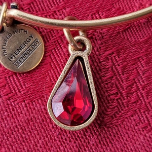Könnte beinhalten: Goldfarbenes Armband mit einem tropfenförmigen, facettierten roten Anhänger. Der Anhänger ist goldfarben umrandet und hängt am Armband. Ein kleiner, runder Anhänger mit dem Text "INFUSED WITH ENERGY TECHNOLOGY" ist ebenfalls angebracht.