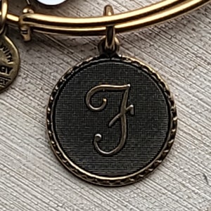 May include: A gold charm bracelet with a black and gold charm featuring the letter "F" in a cursive font.
