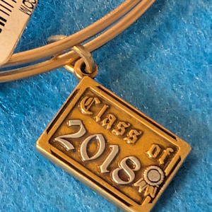 May include: Gold-toned bracelet with a rectangular charm. The charm reads "Class of 2018" in silver lettering. The bracelet has a simple, elegant design, perfect for a graduation gift or a memento of the year.