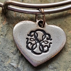 Könnte beinhalten: Silbernes Charm-Armband mit einem herzförmigen Anhänger mit einem monogrammierten Initial. Der Anhänger ist an einem silbernen Armreif befestigt.