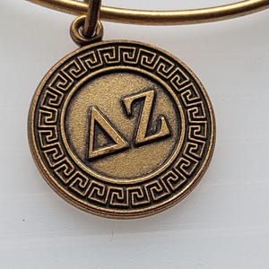 Puede incluir: Una pulsera de metal dorado con un dije circular. El dije presenta un borde de llave griega y las letras griegas Delta y Zeta. La pulsera tiene un diseño simple y elegante, adecuado para el uso diario.