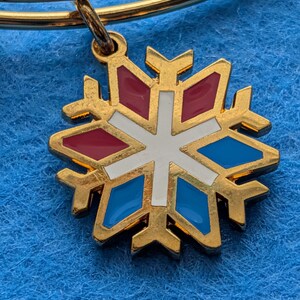 Op de afbeelding: Goudkleurige armband met een sneeuwvlokbedel. De bedel heeft een gouden omtrek met rode, witte en blauwe emaille accenten. De armband wordt gepresenteerd op een blauw, getextureerd oppervlak.