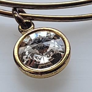 Puede incluir: Primer plano de una pulsera de brazalete dorada con un colgante redondo de cristal transparente. El colgante está engastado en un bisel dorado y cuelga de un pequeño lazo. La pulsera tiene un diseño sencillo y elegante.