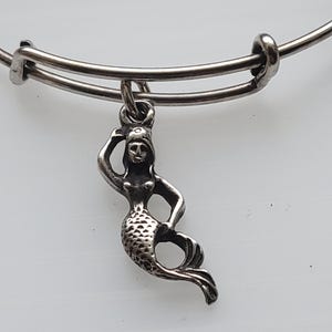 Puede incluir: Pulsera con dije de sirena plateada. El dije es una figura detallada de sirena con cola escamada y cabello suelto. La pulsera tiene un diseño simple y ajustable con un cierre deslizante.