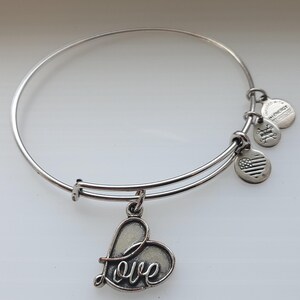 May include: Silver-toned bangle bracelet with a heart-shaped charm that says "Love". The bracelet has a simple, elegant design with several small, round charms and a charm with the American flag.