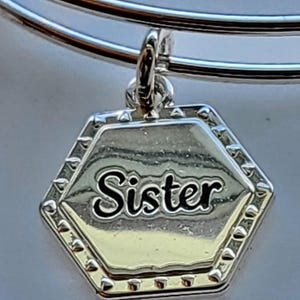 Puede incluir: Pulsera de brazalete plateada con un dije hexagonal. El dije presenta la palabra "Sister" en escritura negra. El dije tiene un borde plateado con pequeños detalles en relieve.