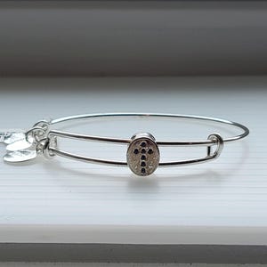 Op de afbeelding: Een zilverkleurige armband met een kruisvormige bedel. De ovale bedel heeft een getextureerd oppervlak en een kruisontwerp gemaakt van kleine, donkere stenen. Extra bedels hangen aan de armband.