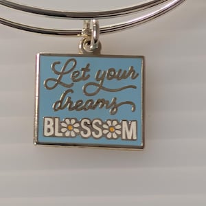 Puede incluir: Pulsera de brazalete plateada con un dije cuadrado. El dije tiene un fondo azul claro con las palabras "Let your dreams BLOSSOM" en letras doradas. La palabra "BLOSSOM" tiene acentos de margarita.