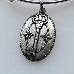 Puede incluir: Charm de pulsera plateado con un colgante ovalado con un diseño de llave. La llave está centrada, rodeada de elementos decorativos y un borde de cuentas. El charm está unido a una pulsera de brazalete plateada.