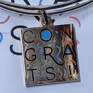 Puede incluir: Pulsera de plata con un dije de plata que dice "CONGRATS" en letras negras con acentos azules y rosas.