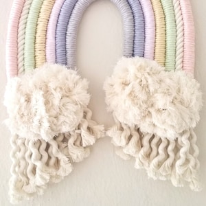 May include: A pastel rainbow wall hanging made of macrame with fluffy white clouds and tassels. The rainbow is made of multiple strands of rope in shades of pink, yellow, green, and purple. The tag attached to the rainbow reads "fi town atelier".