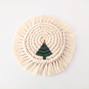 May include: A round, white crocheted coaster with a green and orange Christmas tree design in the center. The coaster has a fringe border.