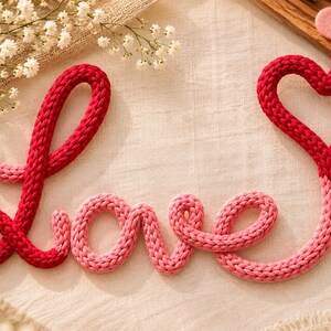 May include: A knitted cord spelling the word love in cursive script, featuring a gradient of red and pink yarn. The letter e is shaped into a heart at the top. Several small, flat felt hearts in shades of red and pink are scattered nearby.