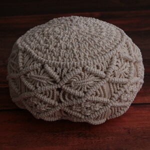 May include: A cream-colored, round, crocheted pouf with a textured surface. The pouf is made of thick, woven cotton rope.