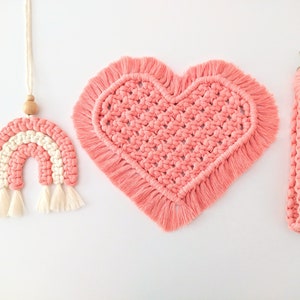 Könnte beinhalten: Ein Set aus drei rosa und weißen Makramee-Artikeln: ein herzförmiger Wandbehang, ein regenbogenförmiger Wandbehang und ein langer, dünner Riemen.