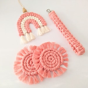 Könnte beinhalten: Ein Set aus vier rosa Makramee-Artikeln: ein Regenbogen-Wandbehang, zwei runde Untersetzer und ein langer Riemen mit einem Metallring. Der Regenbogen hat weiße Streifen und weiße Quasten. Die Untersetzer haben Fransen an den Rändern.
