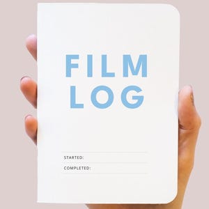 May include: A white notebook with rounded corners featuring the text FILM LOG in light blue capital letters. Below the title, there are two horizontal lines with the labels STARTED: and COMPLETED: printed in small black sans-serif font.