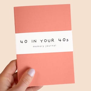 May include: A coral memory journal with the text "40 IN YOUR 40s" and "memory journal" printed on a white band. The journal is held by a hand with light pink nail polish.