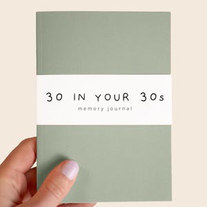 May include: A sage green journal with a white paper band wrapped around the centre. The band features the black printed text 30 IN YOUR 30s and the smaller text memory journal centred below it in a simple, clean, sans-serif typeface.