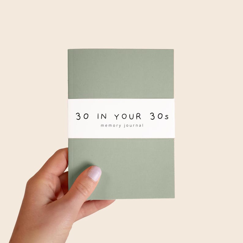 May include: A sage green journal with a white paper band wrapped around the center. The band features the black text 30 IN YOUR 30s and the word memory journal printed in a smaller font below the main title on the front cover.