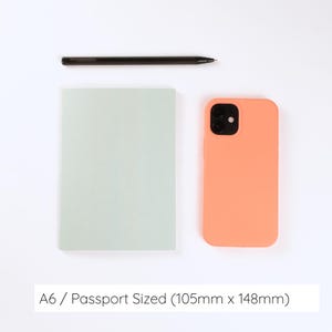 May include: A light green A6 notebook measuring 105mm x 148mm, a black pen, and a peach-colored smartphone case with a dual-camera cutout. The text A6 / Passport Sized (105mm x 148mm) is printed at the bottom of the image for size reference.