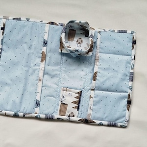 Puede incluir: Un organizador de tela acolchada azul claro con un patrón forestal caprichoso. El organizador presenta un pequeño recipiente redondo y un bolsillo rectangular, ambos adornados con la misma tela con temática forestal. El diseño incluye un patrón de puntos y un borde contrastante.