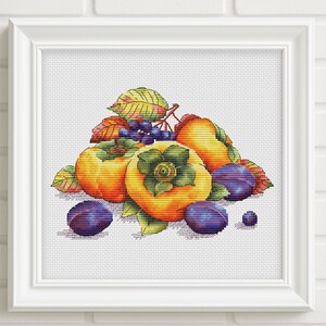May include: A framed cross-stitch embroidery featuring a vibrant still life of orange persimmons, deep purple plums, and small dark berries with green leaves. The artwork is displayed in a square white frame against a white brick wall background.
