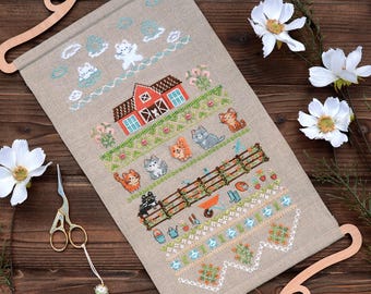 May include: A cross-stitch wall hanging featuring a pastoral scene with cats, a red barn, and a wooden fence. The design includes various colors and floral elements, displayed on a beige fabric. The hanging is mounted on a decorative wooden frame.