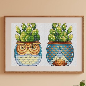 May include: Cross-stitch art featuring two potted cacti. One pot is an owl design with blue, yellow, and gold accents. The other pot has a blue and orange geometric pattern. The cacti are green with white dots.