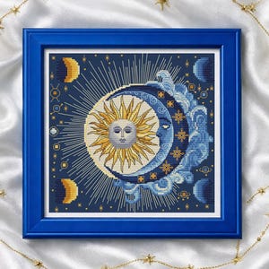 May include: A framed cross-stitch artwork featuring a celestial design with a sun and moon motif in shades of yellow and blue. The artwork is in a blue frame, with gold star-shaped embellishments.