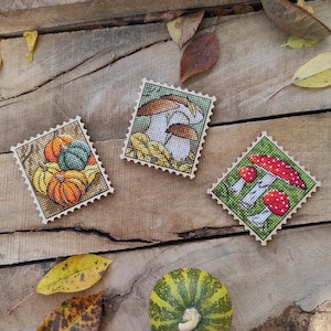 Puede incluir: Tres cuadrados de madera con diseños de punto de cruz de calabazas, setas y una seta con puntos rojos. Los cuadrados están sobre una superficie de madera con hojas de otoño.