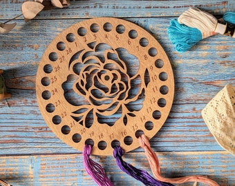 May include: A wooden thread organizer with 28 holes, featuring a carved rose design. The organizer has a light brown color and is surrounded by colorful embroidery floss.