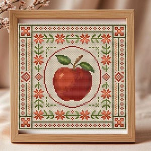 Op de afbeelding: Een ingelijst kruissteekkunstwerk met een rode appel met groene bladeren, gecentreerd in een cirkelvormig ontwerp. Het kunstwerk is omgeven door een rand van oranje bloemen en groen blad, alles tegen een crèmekleurige achtergrond. Het frame is gemaakt van lichtgekleurd hout.