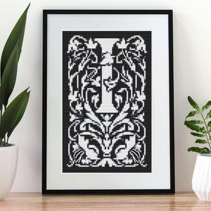 Op de afbeelding: Zwart omlijst kruissteekkunstwerk met de witte letter "I", omgeven door een sierlijk bloemmotief op een zwarte achtergrond. Het kunstwerk wordt getoond op een houten oppervlak met potplanten op de achtergrond.
