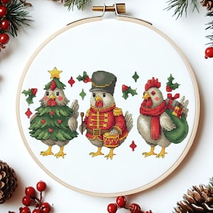 Può includere: Un telaio da ricamo a punto croce con tre galline festive. Una è vestita da albero di Natale, un'altra da tamburino in uniforme rossa, e la terza indossa una sciarpa e porta una borsa regalo. Il disegno è circondato da aghi di pino, bacche rosse e pigne.