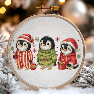 May include: A cross-stitch embroidery hoop featuring three festive penguins. One penguin wears red and white striped pajamas and a hat, holding a mug. Another is in a green sweater with lights, and the third wears a red hoodie and hat, holding a gift. Snowflakes and Christmas decor surround the hoop.