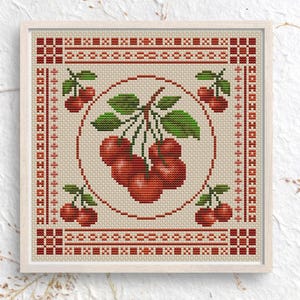 May include: Cross-stitch artwork featuring a cluster of red cherries with green leaves in a circular design. The artwork is framed in a light-colored wooden frame, with a decorative border in red and cream colors. The piece has a folk art aesthetic.