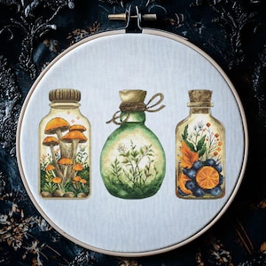 Könnte beinhalten: Kreuzstich-Stickerei mit drei Glasflaschen mit Korken, die jeweils mit verschiedenen botanischen Elementen gefüllt sind. Die Flaschen werden in einem hölzernen Stickrahmen präsentiert. Die erste Flasche enthält Pilze, die zweite Grünzeug und die dritte Beeren und Orangenscheiben.