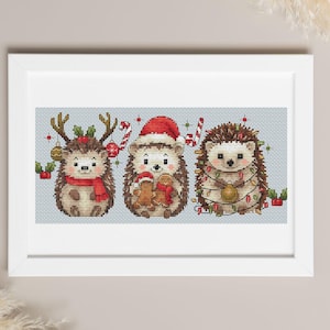 May include: A framed cross-stitch featuring three festive hedgehogs. The hedgehogs are decorated with Christmas elements like reindeer antlers, Santa hats, candy canes, and string lights. The artwork is set against a light blue background.
