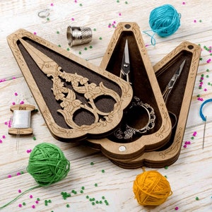 May include: A wooden box with three compartments, each containing a pair of decorative scissors. The box is intricately carved with floral designs and has a dark brown stain. The scissors are silver and have ornate handles.