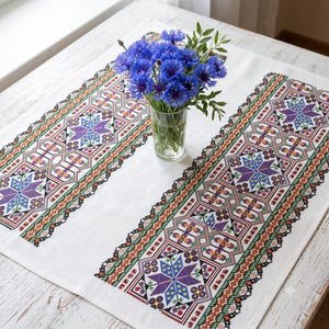May include: A white rectangular table runner featuring two wide vertical bands of intricate cross-stitch embroidery in purple, green, red, and black, displaying geometric star patterns. A clear glass vase holds a bouquet of blue cornflowers.