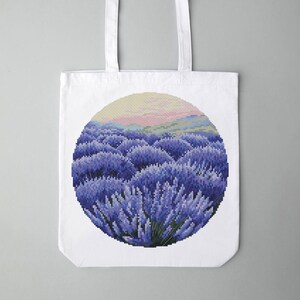 May include: White tote bag featuring a circular design of a lavender field. The artwork depicts rows of purple lavender plants under a yellow and pink sky. The bag has two white handles.