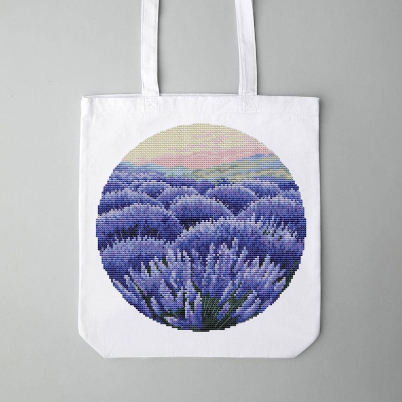 May include: White tote bag featuring a circular design of a lavender field. The artwork depicts rows of purple lavender plants under a yellow and pink sky. The bag has two white handles.