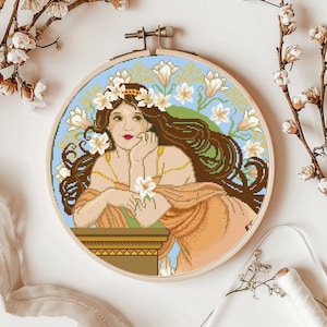 May include: A cross-stitch embroidery hoop with a colorful image of a woman with long brown hair wearing a crown of white flowers. The woman is surrounded by white flowers and a blue sky.