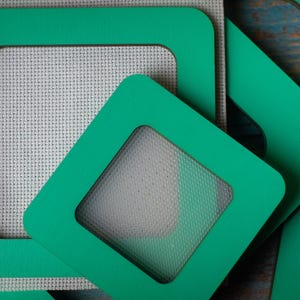 Op de afbeelding: Close-up van verschillende vierkante, teal-gekleurde frames met een vierkante uitsparing. De frames zijn gestapeld op een stuk witte kruissteekstof. De frames hebben een gladde, matte afwerking en zijn in verschillende hoeken gerangschikt.