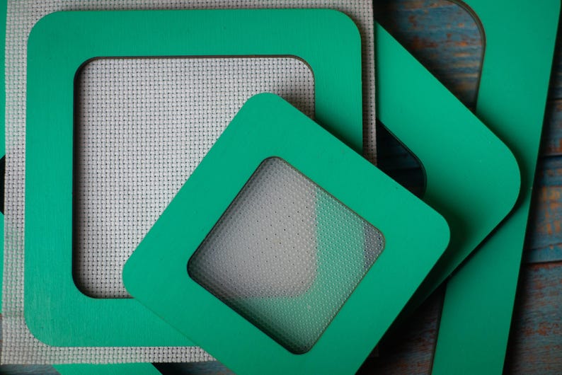 Op de afbeelding: Close-up van verschillende vierkante, teal-gekleurde frames met een vierkante uitsparing. De frames zijn gestapeld op een stuk witte kruissteekstof. De frames hebben een gladde, matte afwerking en zijn in verschillende hoeken gerangschikt.