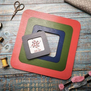 Op de afbeelding: Een verzameling knutselspullen, waaronder vier vierkante houten frames in rood, groen, blauw en grijs. Het kleinste frame bevat een kruissteek met het woord "DREAM". Ook aanwezig zijn scharen, draad en spoelen.