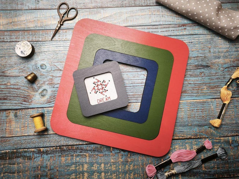 Op de afbeelding: Een verzameling knutselspullen, waaronder vier vierkante houten frames in rood, groen, blauw en grijs. Het kleinste frame bevat een kruissteek met het woord "DREAM". Ook aanwezig zijn scharen, draad en spoelen.